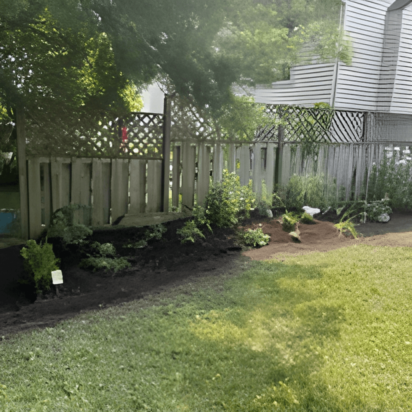Newly planted garden bed with dark soil bordering a wooden fence and green lawn.